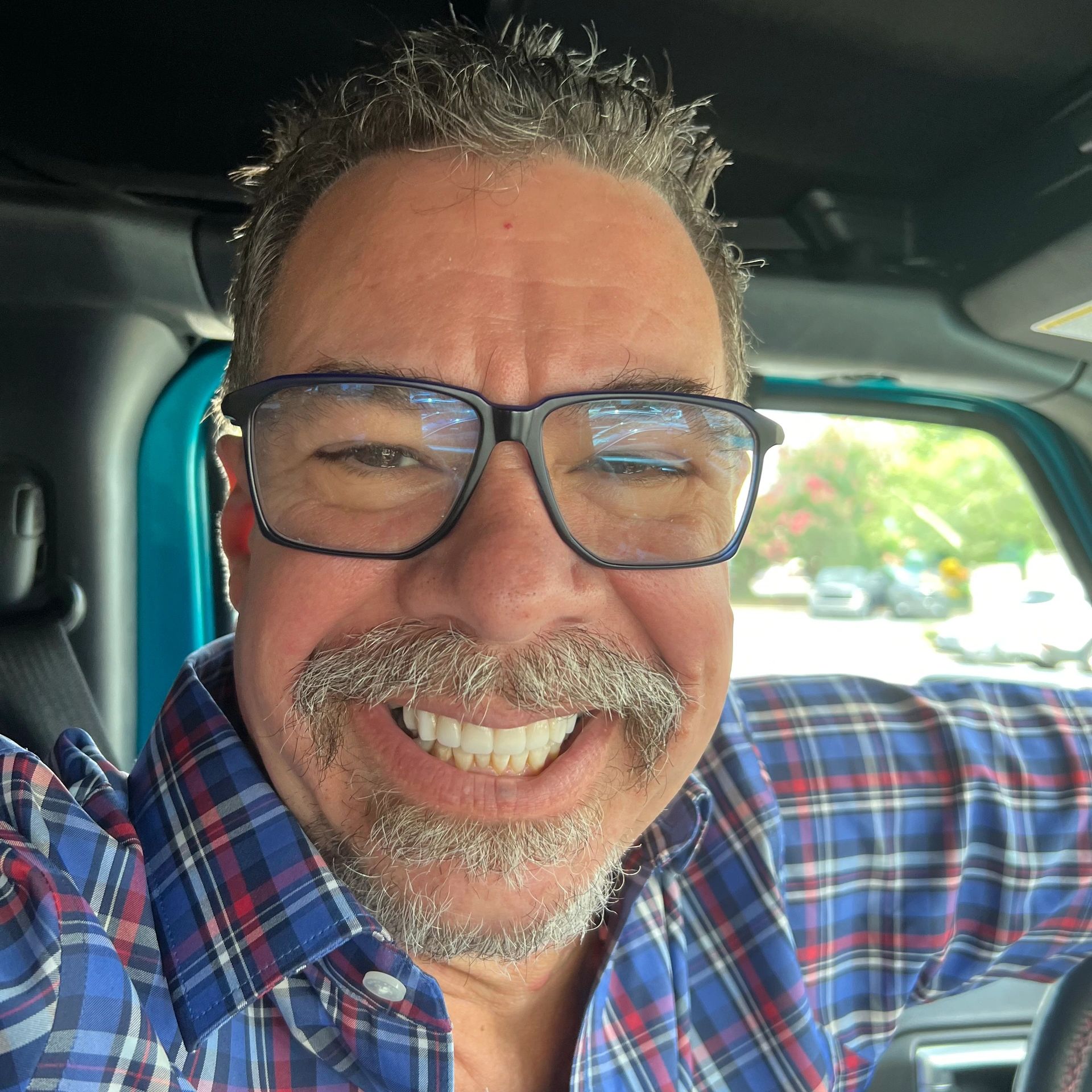 Smiling man with glasses and a mustache wearing a plaid shirt inside a vehicle.