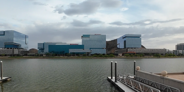 Modern office buildings reflect on a calm lake under a cloudy sky.