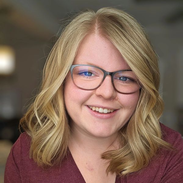 Smiling woman with blonde hair and glasses in a cozy indoor setting.
