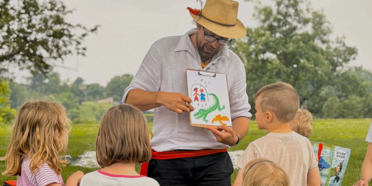 Man in a hat shows a colorful drawing to children outdoors.
