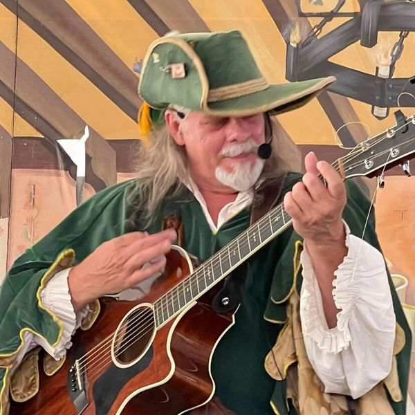 Man in Renaissance attire playing acoustic guitar with headset mic.