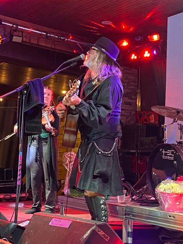 Musician playing guitar and singing on stage with violinist.