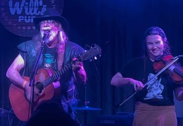 Two musicians perform on stage, one playing guitar and singing, the other playing violin.