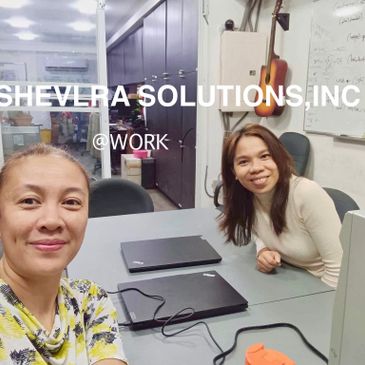 Two women smiling at a workspace with laptops and office equipment.
