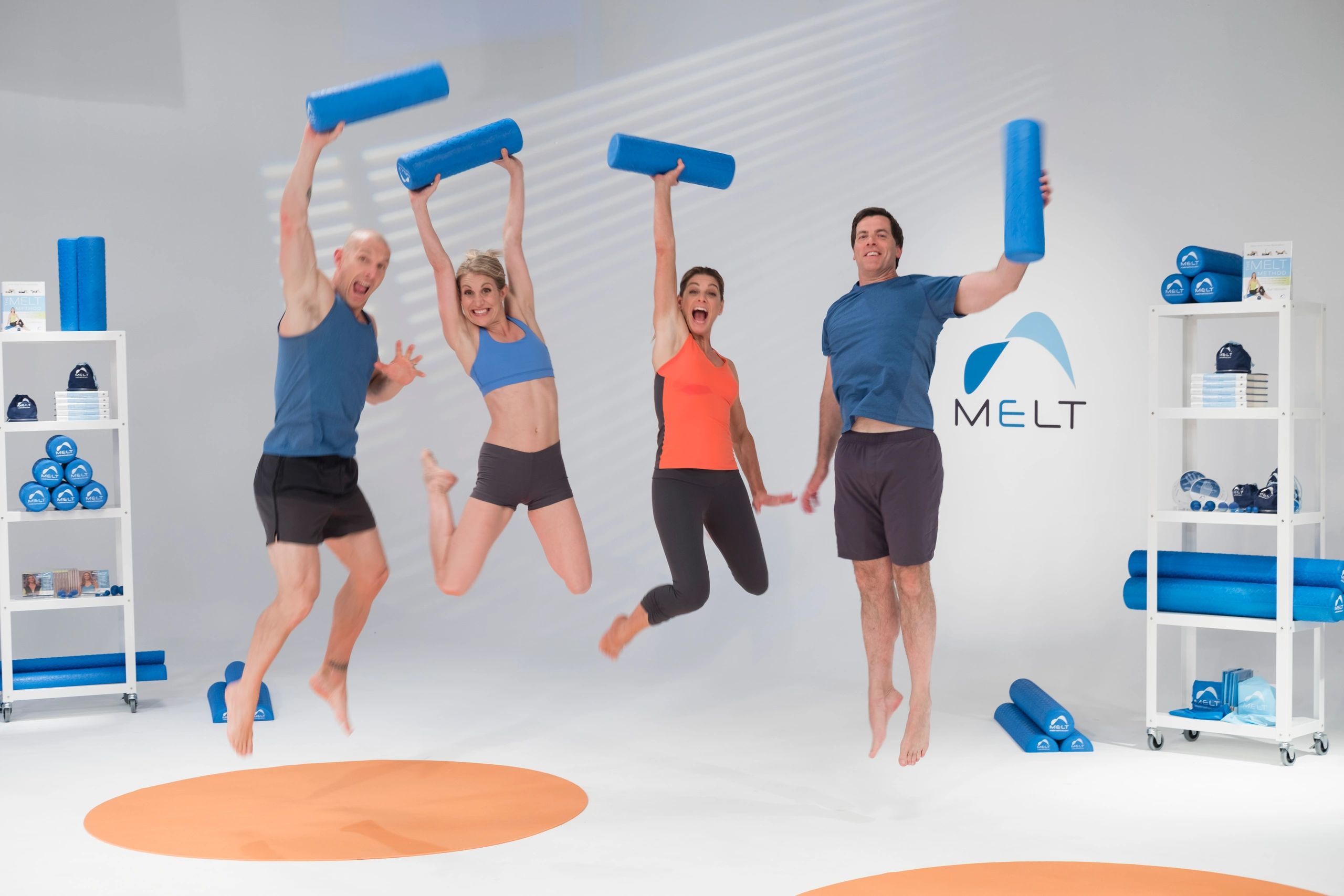 Four adults jumping holding blue foam rollers in a fitness studio.
