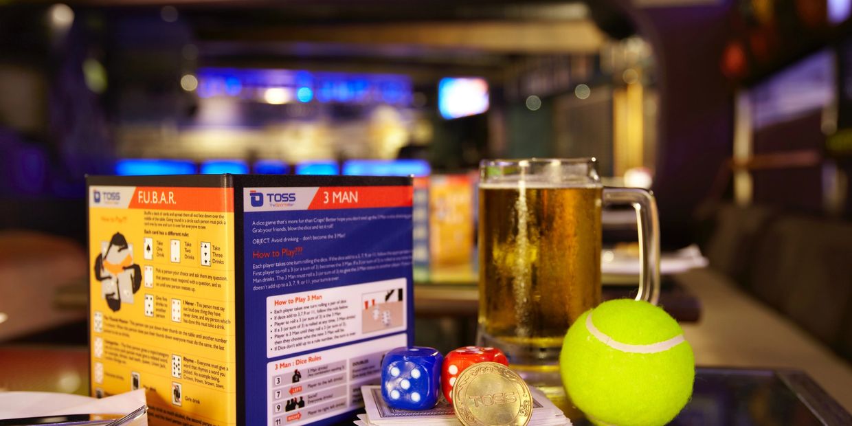 A glass of beer, tennis ball, dice, and game instructions on a bar table.