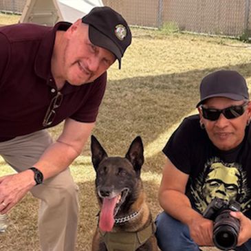 Two men posing with a happy dog outdoors on a sunny day.