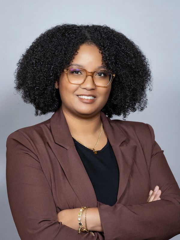 Confident woman with curly hair and glasses wearing a brown blazer.