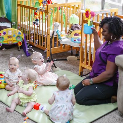 Teacher blows bubbles with babies in a classroom setting. Cribs for infant care in a large classroom