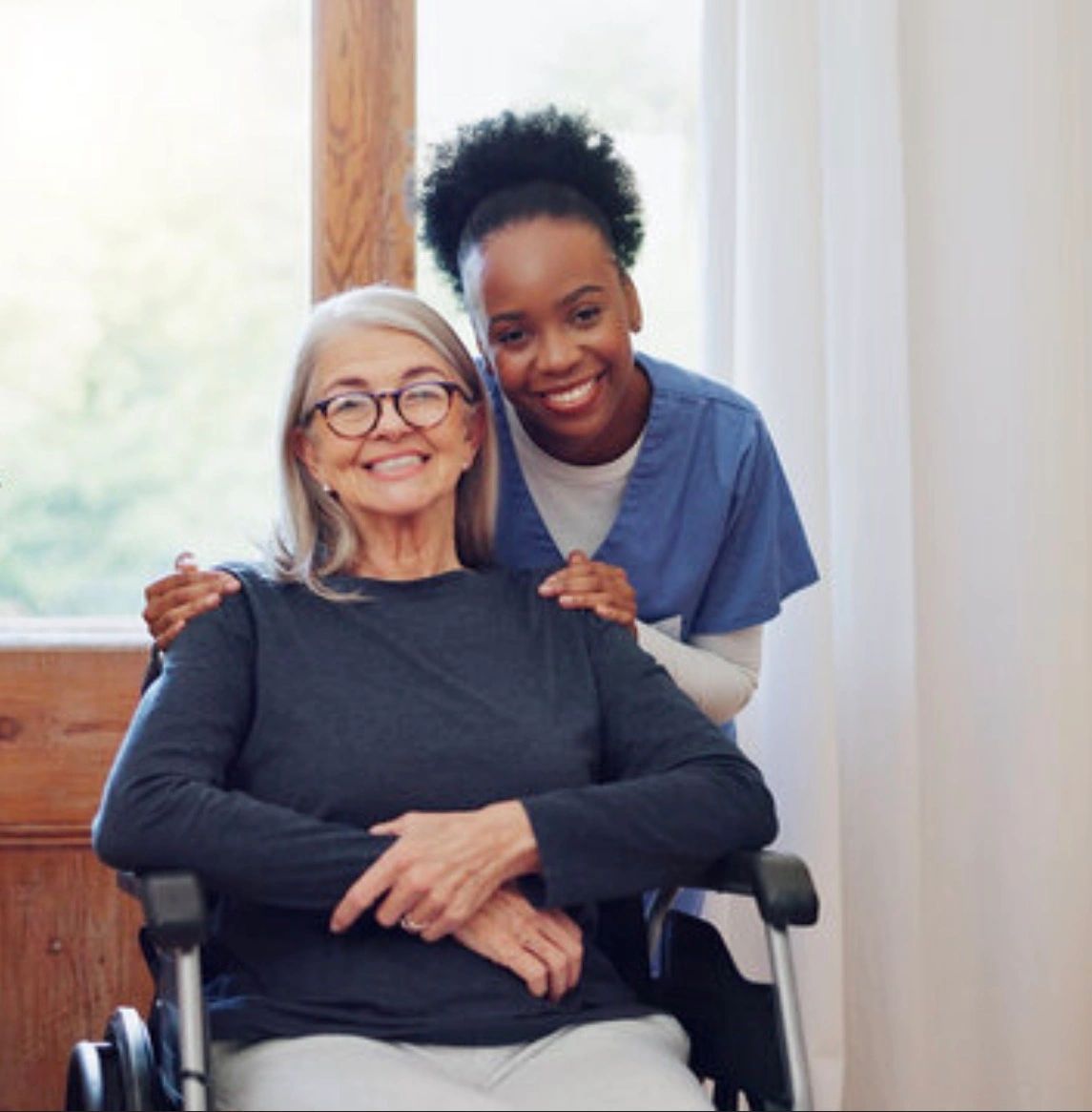 Smiling elderly woman in wheelchair with a caring nurse behind her.