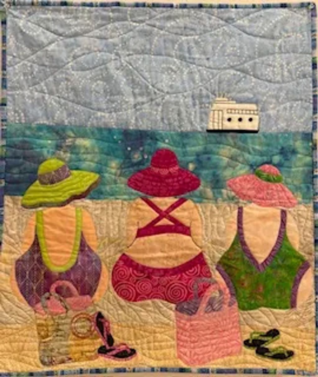 Quilt art of three women in colorful swimsuits and hats at the beach, with a boat in the distance.