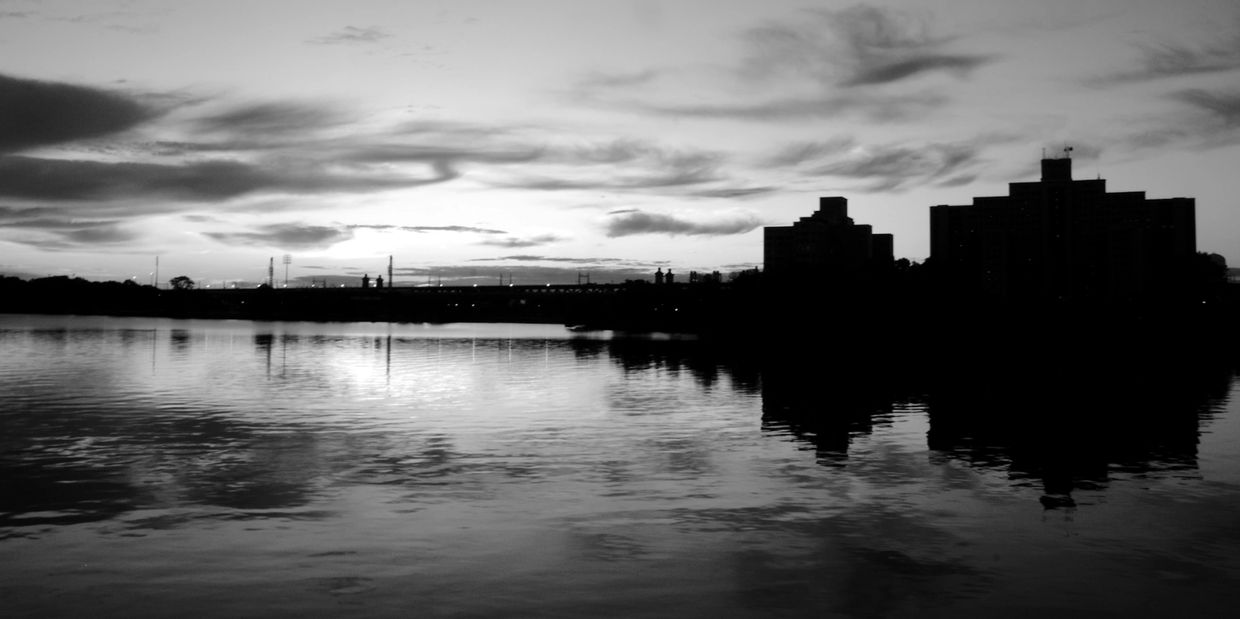 Black and white cityscape with buildings reflected in calm water at dusk.