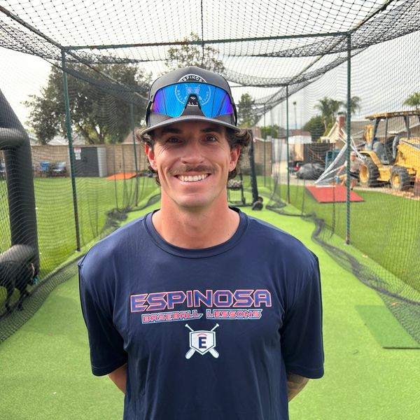 A baseball coach stands smiling in a batting cage wearing an Espinosa Baseball Lessons shirt.