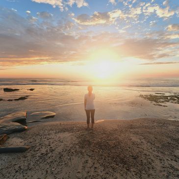 A person stands on a beach watching a glowing sunset over the ocean.
