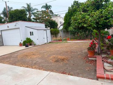 A bare backyard with a small tree and potted plants near a white garage.