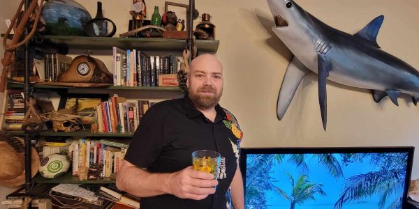 Man holding a drink in a room with bookshelf and mounted shark.