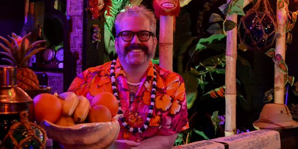 Man in a vibrant Hawaiian shirt smiling behind a tiki bar.