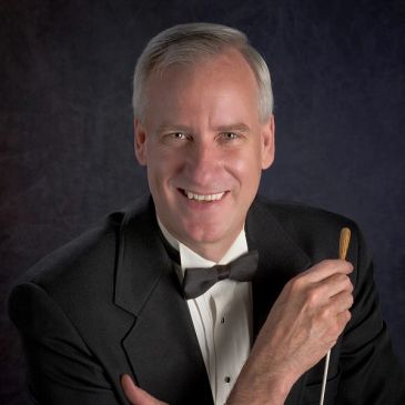 A smiling man in a tuxedo holding a conductor's baton.