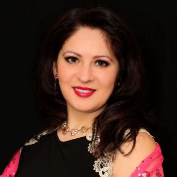 A woman with dark hair wearing a black dress and pink shawl smiles against a black background.