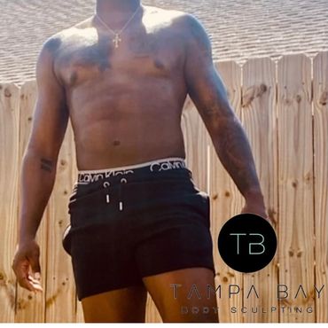 Shirtless man in black shorts posing outdoors against a wooden fence.