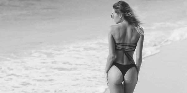 A woman in a bikini stands on a sandy beach looking at the ocean.