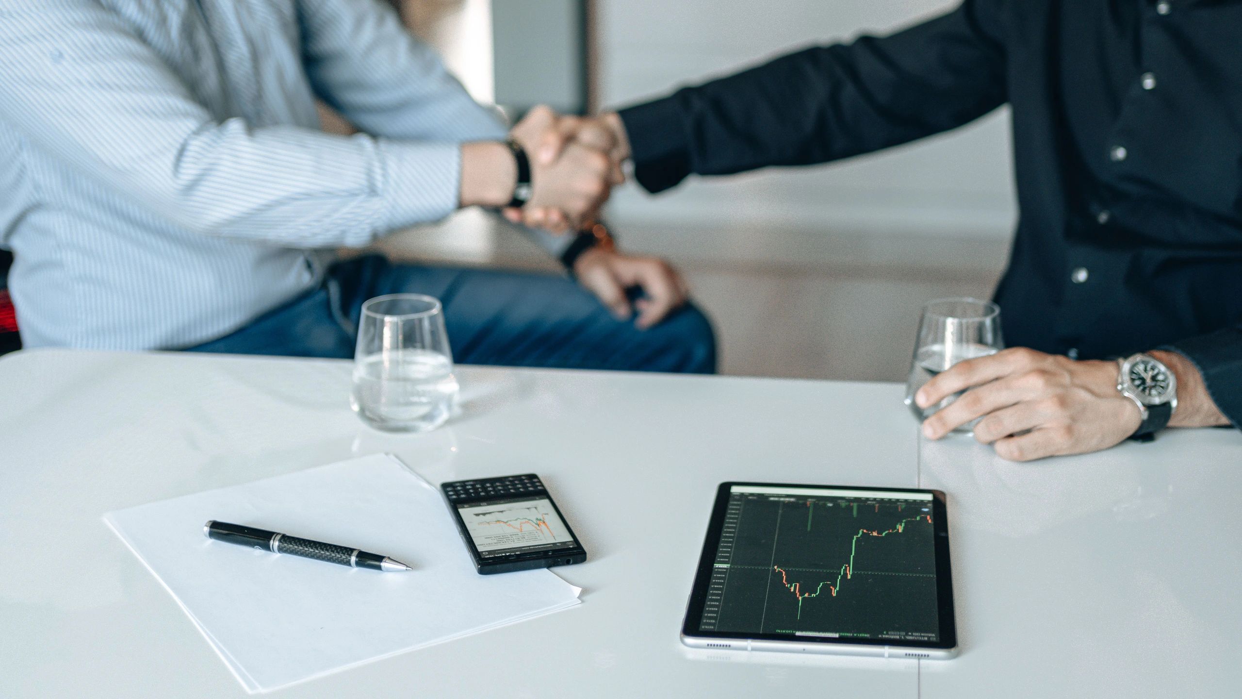 Two people shaking hands over a table with devices and documents.