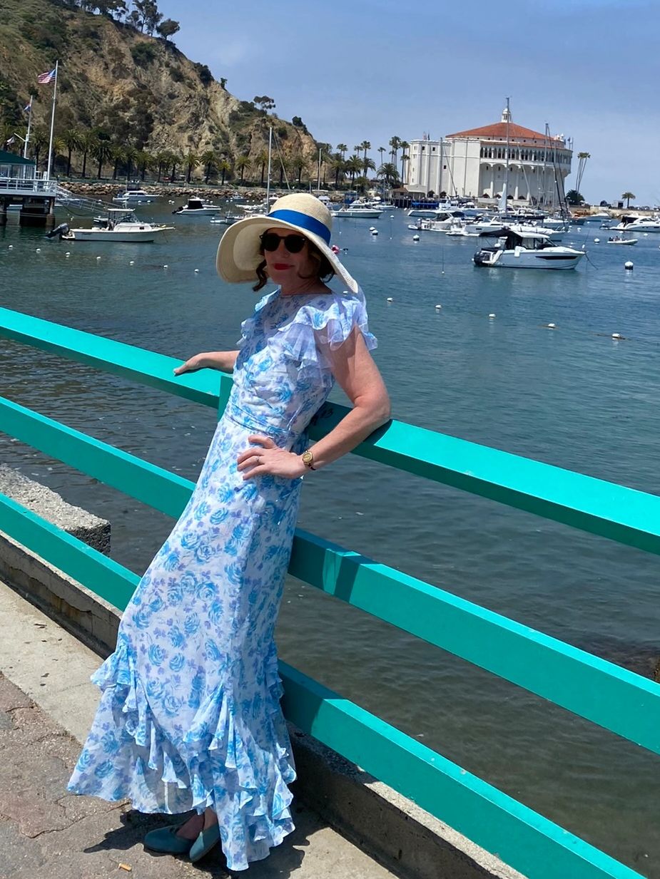 Woman in 1930s-style blue floral dress & straw hat Catalina Island harbor with Casino and sailboats.