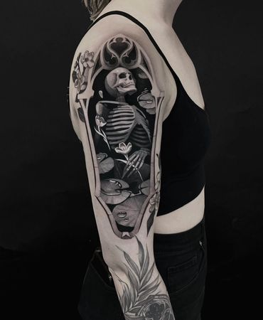 Black and grey tattoo of a skeleton surrounded by flowers on an upper arm.