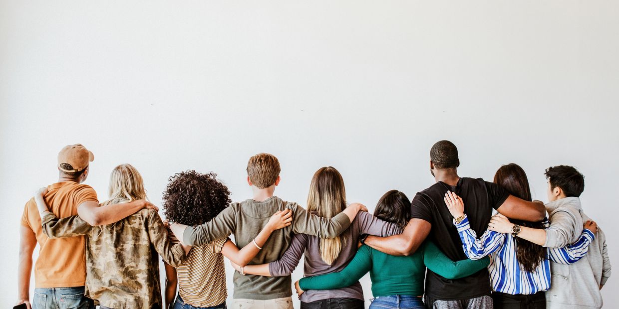 A diverse group of friends stand arm in arm facing a white wall.