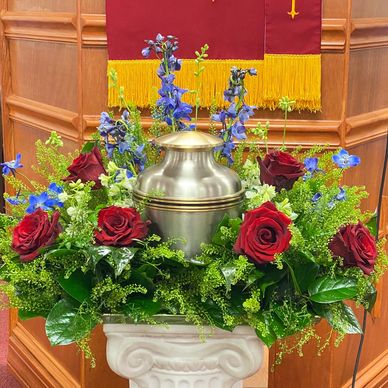 Serene urn surrounded by red roses, dark blue delphinium, Lepidium and lush greens.