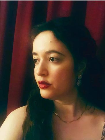 Woman with dark hair looking thoughtfully to the side against a red curtain backdrop.
