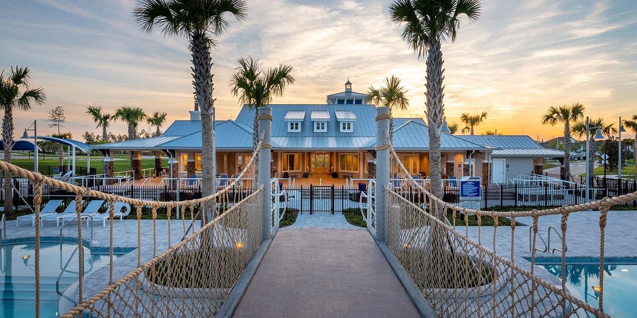 Suspension bridge leading to a clubhouse at sunset with palm trees and pools.