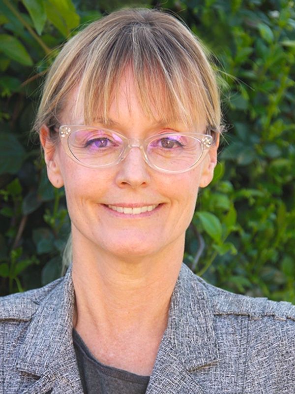 Smiling woman wearing clear glasses and a gray blazer outdoors.