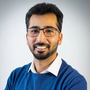Smiling man with glasses and beard wearing a blue sweater over a white shirt.