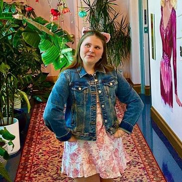 A young woman poses in a colorful hallway with hanging flowers and green plants.