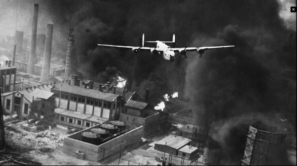 Bomber flying over smoking building.
