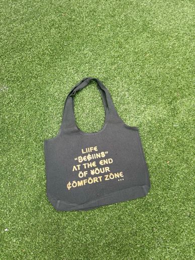 Black tote bag with motivational quote on green grass.