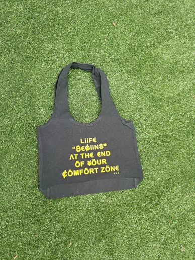 Black tote bag with motivational yellow text on green grass.