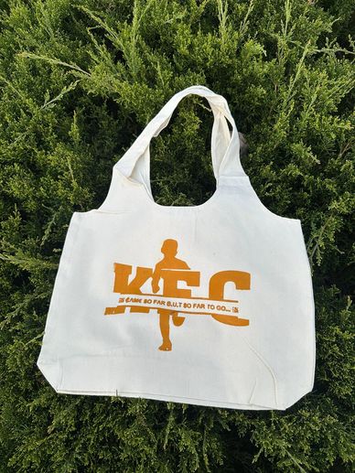 White tote bag with orange running-themed print on green foliage background.