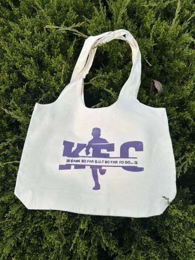 White tote bag with a purple KFC silhouette design on green foliage.