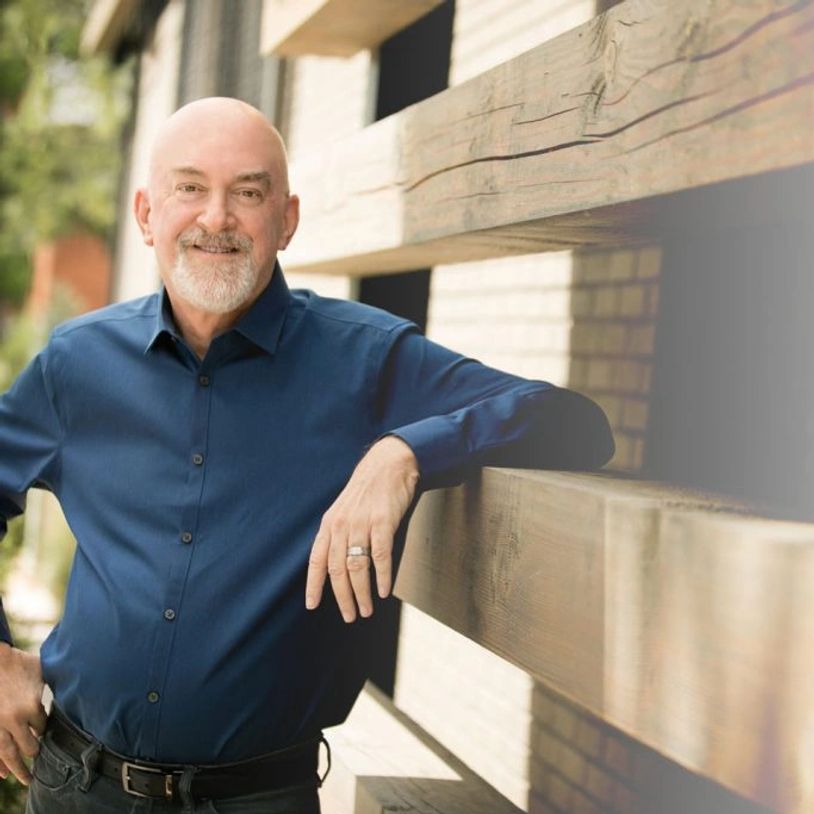 Smiling bald man in a blue shirt leaning on a wooden structure outdoors.