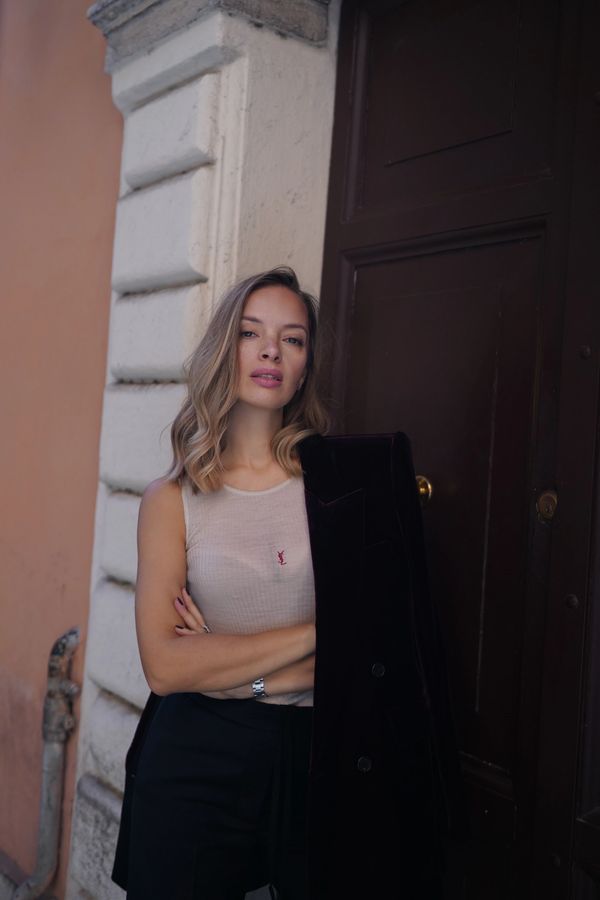 Woman standing by a doorway with arms crossed, wearing a sleeveless top and holding a jacket.