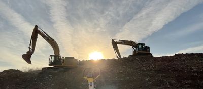 Two excavators work on a dirt mound during sunset with a partly cloudy sky.