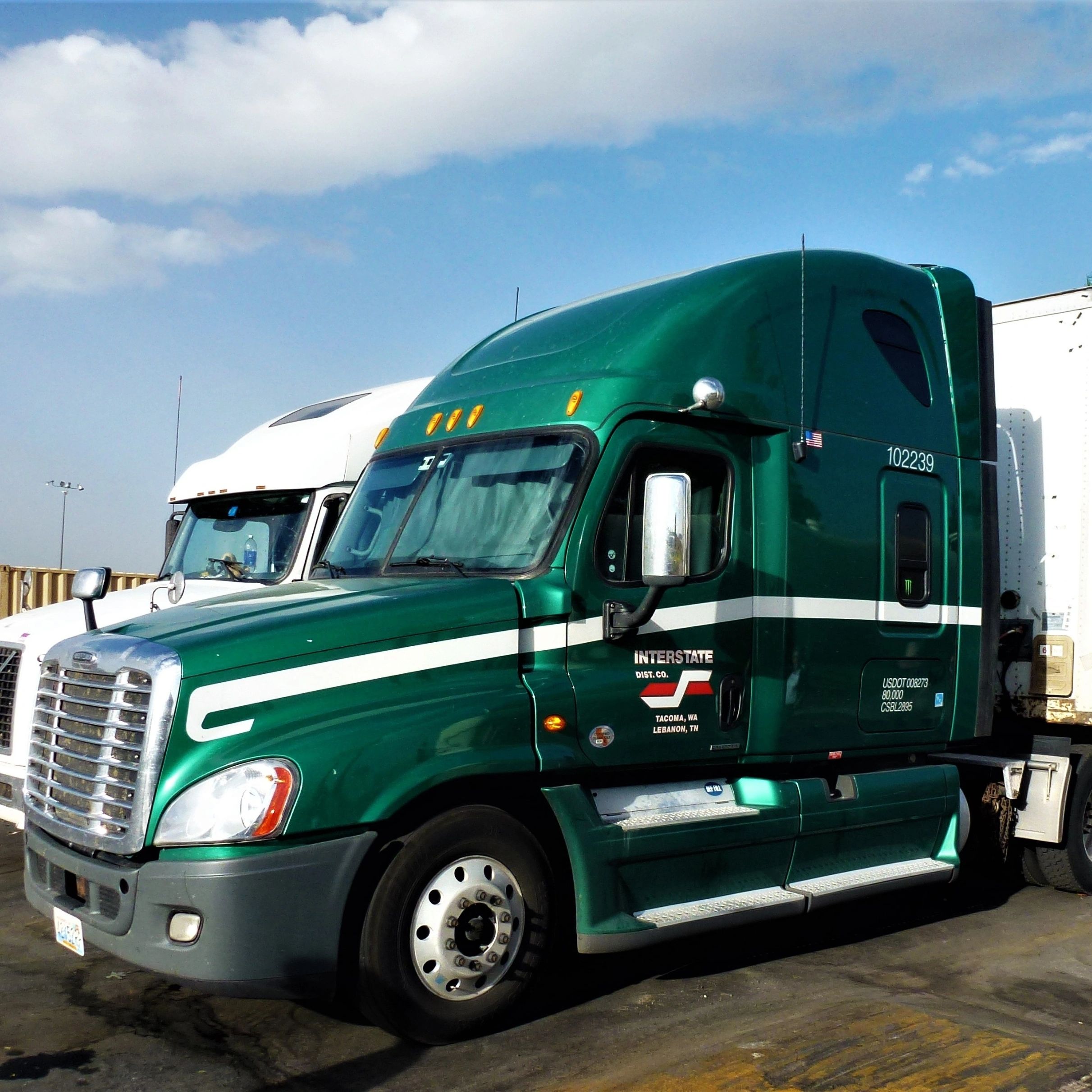 Green semi truck parked beside a white truck under a partly cloudy sky.