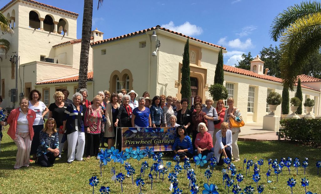 Group of women posing in front of a building with blue pinwheels on the lawn.