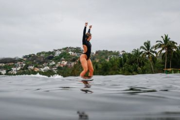 Woman in black swimsuit standing with arms raised in water near a tropical shoreline.