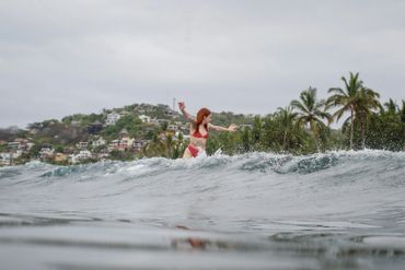 A woman in a red bikini surfing a wave near a tropical coastline.