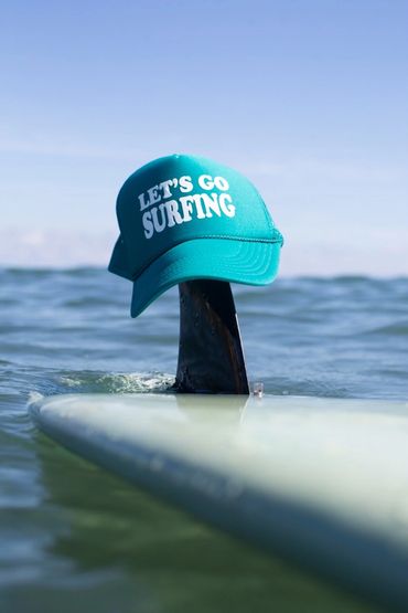 A teal cap with 'LET'S GO SURFING' on a surfboard fin in the ocean.