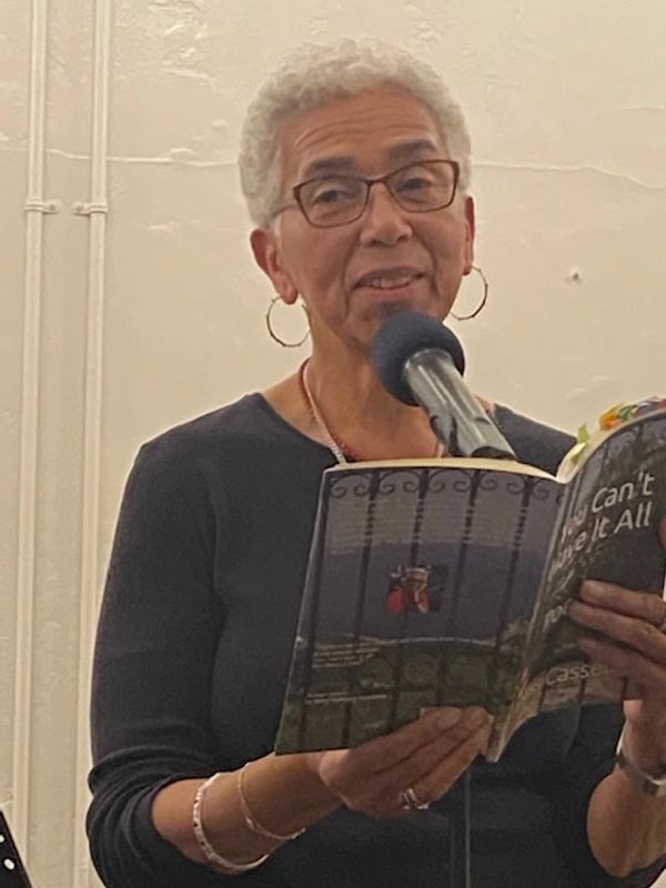 Black woman with white hair standing at a microphone, dressed in a black shirt, holding and reading 