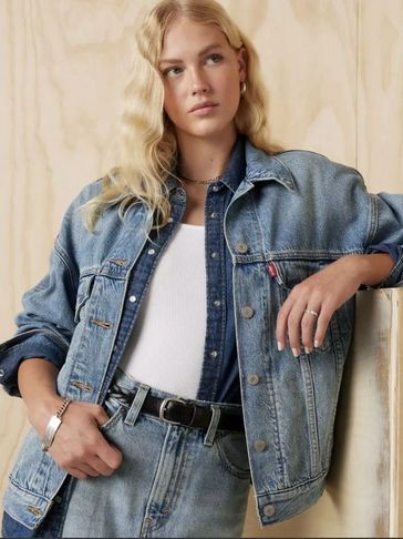 Young woman in layered denim jacket and jeans poses against a light wooden background.